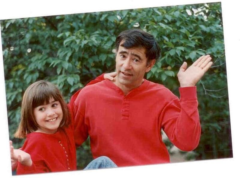 A man and a girl smiling outdoors with greenery.