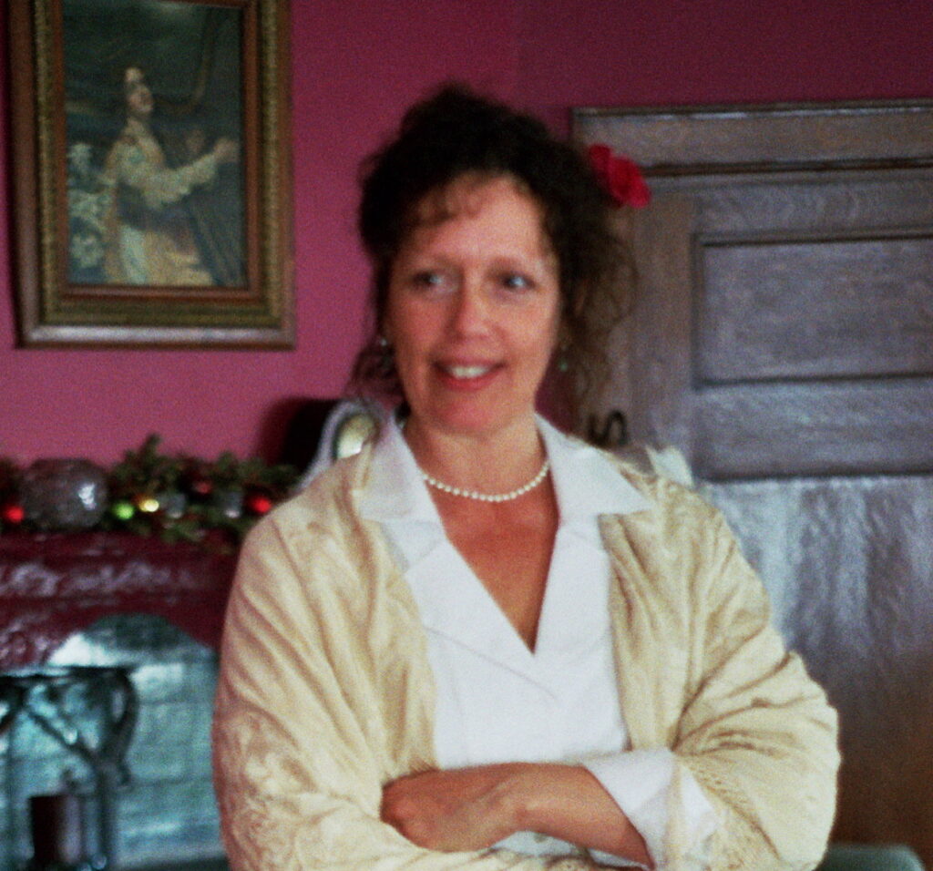 A smiling woman in a beige shawl standing indoors.
