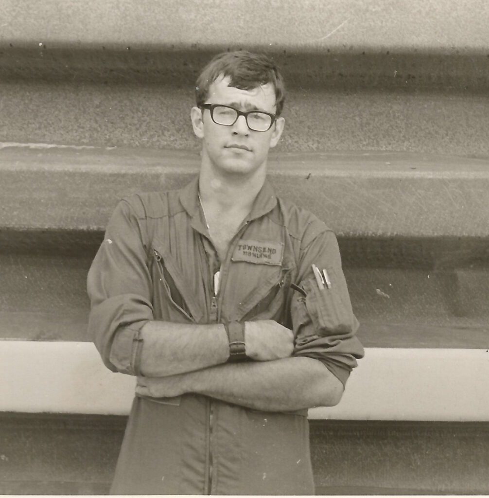 Young man in glasses and workwear standing indoors with arms crossed.