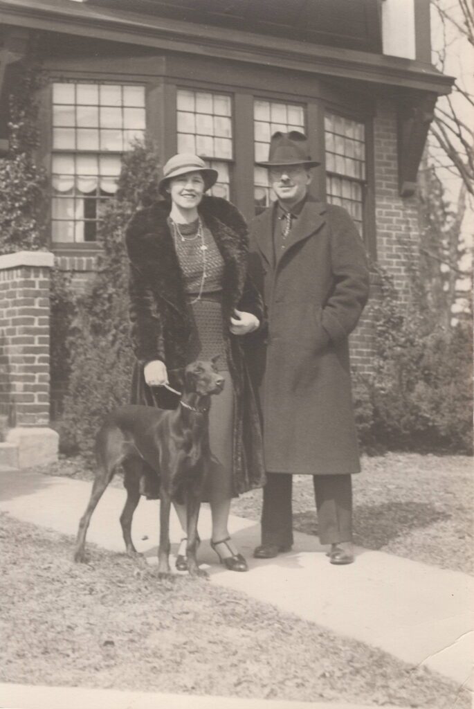 Vintage photo of a man and woman standing with a dog outside a brick house.