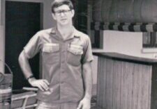 A young man in glasses and a uniform stands confidently indoors.