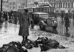 A man in a coat stands near bodies on a wet city street during wartime.