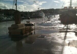 A harbor scene with boats docked under a bright sky and wet concrete reflecting sunlight.